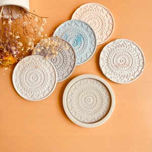 Può includere: Una collezione di sottobicchieri rotondi decorativi in vari colori, tra cui bianco, azzurro e beige. Ogni sottobicchiere presenta un design mandala in rilievo. I sottobicchieri sono disposti su una superficie arancione chiaro, con fiori secchi sullo sfondo.