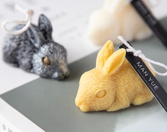 May include: Two small rabbit-shaped candles, one is gray and the other is yellow. The yellow candle has a tag that reads "MAN YUE THE CREATION OF FLOWERS AND FRAGRANCE".