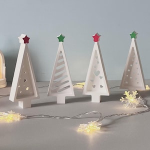 May include: Four white Christmas tree ornaments with red and green star toppers. The trees feature cut-out designs of hearts, lines, and circles. A string of snowflake fairy lights is in front, creating a festive holiday display.
