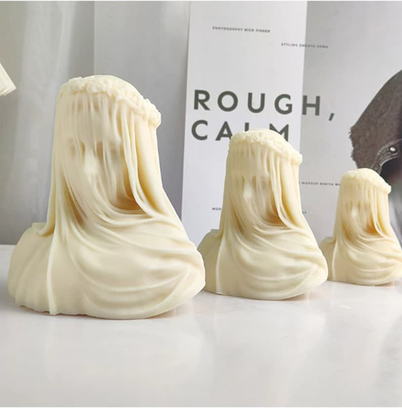 May include: Three off-white sculptures of veiled busts. The sculptures are arranged in a row, decreasing in size from left to right. The busts have a draped fabric effect, with the veil cascading down the front. The background includes a white surface and a poster with the words "ROUGH, CALM."
