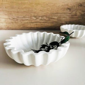 Op de afbeelding: Een witte keramische kom met een gegolfde rand. De kom is leeg en heeft een takje met groene bladeren erin.