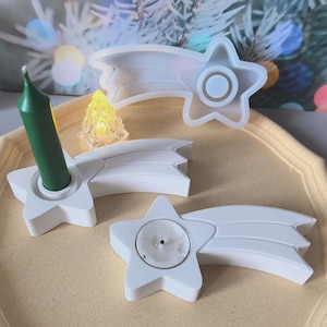 Puede incluir: Candelabros blancos en forma de estrella y un molde de silicona para manualidades. Un soporte tiene una vela verde. Los soportes tienen un diseño de cola de cometa. Una pequeña luz amarilla en forma de árbol de Navidad de cristal está en el fondo.