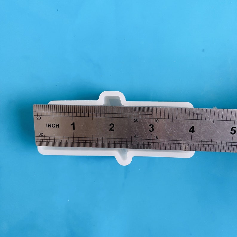 Op de afbeelding: Een wit rechthoekig object met een liniaal dat een lengte van ongeveer 10 cm aangeeft. Het object heeft een unieke vorm met twee uitstekende delen aan weerszijden. De achtergrond is effen lichtblauw.