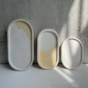 May include: Three oval-shaped concrete trays in different sizes. The largest tray has a light yellow and white marbled design. The two smaller trays are solid white.