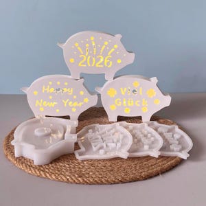 Puede incluir: Un conjunto de decoraciones en forma de cerdo gris claro con detalles y texto amarillos. Un cerdo dice "2026", otro dice "Happy New Year" y un tercero dice "Viel Glück". Las decoraciones están sobre una estera tejida.