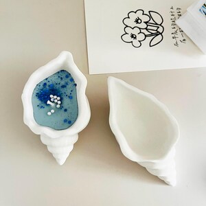 May include: Two white seashell-shaped candle holders. One holder has a blue candle with white and blue glitter inside.