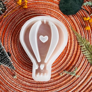 Puede incluir: Molde de silicona blanco con forma de globo aerostático con un pequeño corazón en el centro. El molde está sobre una superficie circular marrón y naranja.