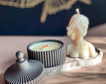 May include: A black and white candle holder with a lid and a white candle in the shape of a bust. The candle holder has a ribbed design and the bust has a detailed face.