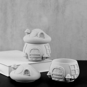May include: Three white ceramic mushroom-shaped containers with a rounded top and a base with a door and windows. The containers are on a black surface.