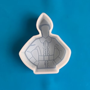 May include: A clear silicone mold in the shape of a cartoon elf with arms akimbo. The elf is wearing a hat, shirt with ruffles, and a belt. The mold is on a blue background.