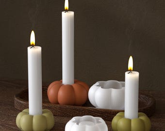 May include: A collection of pumpkin-shaped candle holders in shades of orange, green, and white, each holding a lit white taper candle. The candles are arranged on a wooden tray, creating a warm, autumnal ambiance.