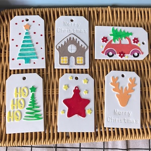 May include: A collection of white Christmas gift tags with festive designs. Each tag features a different holiday image, including a Christmas tree, gingerbread house, car with a tree, and a reindeer. Some tags have the text "Merry Christmas" or "HO HO HO".