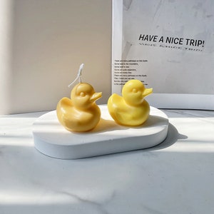 Könnte beinhalten: Zwei gelbe, gummienteförmige Kerzen auf einer weißen, ovalen Oberfläche. Die Kerzen sind aus Wachs und haben ein realistisches Enten-Design.