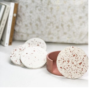 May include: Set of four white and pink terrazzo coasters with a matching terrazzo lidded container. The coasters and container have a speckled pattern of pink and white.