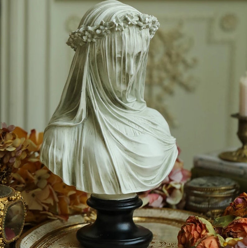 May include: A white bust sculpture of a veiled woman, adorned with a floral crown. The sculpture features a draped veil and is mounted on a black pedestal. The bust is a decorative item, likely for home decor.