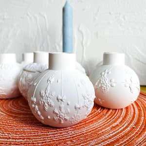 以下が含まれることがあります： エンボス加工が施された白いセラミック製のキャンドルホルダーが5つ。キャンドルホルダーは丸型で、表面にテクスチャがあります。赤と白のストライプのプレースマットの上に並べてあります。