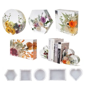 Op de afbeelding: Een assortiment van transparante hars boekenleggers en decoratieve objecten met geperste bloemen en groen. De boekenleggers zijn hartvormig en tonen een verscheidenheid aan bloemen in tinten roze, geel en oranje. De decoratieve objecten zijn in de vorm van een zeshoek, een cirkel en een rechthoek. Alle objecten zijn gemaakt van transparant hars en tonen geperste bloemen en groen in verschillende tinten groen, roze, geel en oranje.