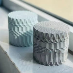 Könnte beinhalten: Zwei dekorative Betonbehälter mit geometrischen Mustern. Der Behälter links ist hellblau und der Behälter rechts ist grau.