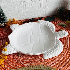 Może przedstawiać: Biały ceramiczny talerz w kształcie żółwia o teksturowanej powierzchni. Talerz ma płytką miseczkę i jest zaprojektowany ze szczegółowymi wzorami płetw i skorupy. Talerz jest umieszczony na pomarańczowo-brązowej okrągłej podkładce.