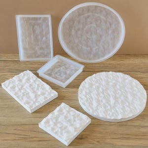 Op de afbeelding: Een assortiment harsmallen en afgewerkte stukken. De mallen zijn transparant en omvatten een rechthoekige bak, een vierkante bak en een grote ronde bak. De afgewerkte stukken zijn wit en getextureerd.