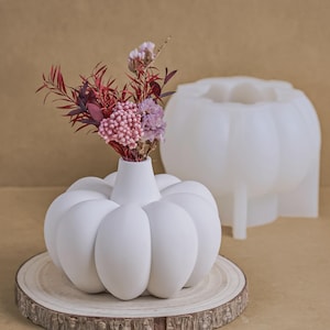 May include: A white pumpkin-shaped vase with a small bouquet of dried flowers in pink and burgundy. The vase is on a wooden slice, with a matching pumpkin mould behind it. The vase has a round base and a narrow neck.