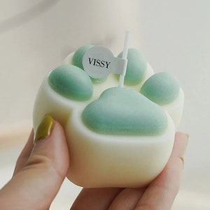 May include: A white paw print shaped candle with green paw pads. The candle has a small white label with the word "VISSY" on it.