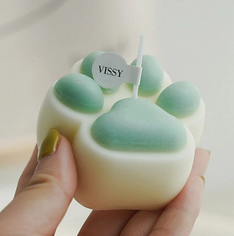 May include: A white paw print shaped candle with green paw pads. The candle has a small white label with the word "VISSY" on it.