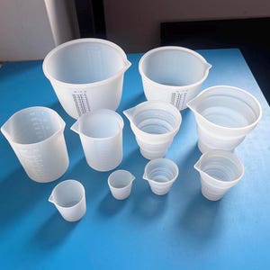 Pode incluir: Um conjunto de nove xícaras medidoras de silicone branco de vários tamanhos. As xícaras são todas transparentes e têm marcas em mililitros e onças.