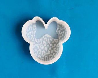 May include: White silicone mold shaped like a cartoon mouse head with a bow. The mold has a leopard print design.
