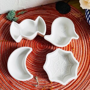 Op de afbeelding: Vier witte keramische Halloween-thema schalenvormen. De vormen zijn in de vorm van een snoepje, een spook, een halve maan en een spinnenweb. De vormen staan op een ronde, rood-witte placemat.