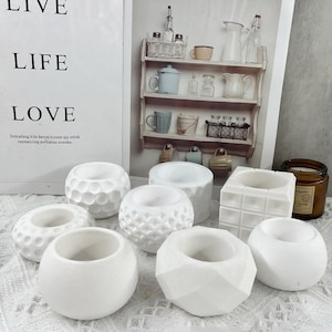 May include: Eight white ceramic candle holders with different geometric designs. The candle holders are arranged on a white lace tablecloth.