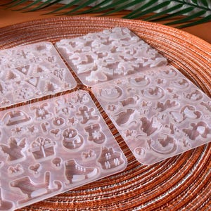 Könnte beinhalten: Vier transparente Silikonformen mit verschiedenen weihnachtlichen Formen. Die Formen umfassen Formen von Weihnachtsbäumen, Sternen, Lebkuchenmännern, Engeln, Geschenken und Schlittschuhen. Die Formen sind auf einem orangefarbenen, gewebten Platzset angeordnet.