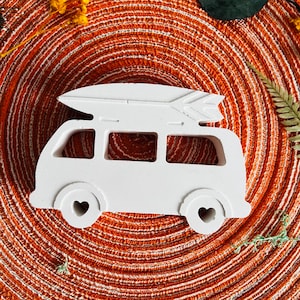 May include: White ceramic figurine of a vintage van with a surfboard on top. The van has heart-shaped wheels and is sitting on a brown woven surface.