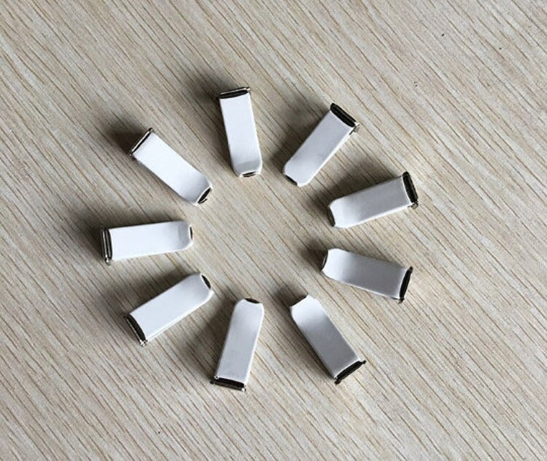 Op de afbeelding: Tien witte plastic clips met een zilveren metalen bevestiging. De clips zijn in een cirkel op een lichtbruin houten oppervlak gerangschikt.