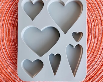 Puede incluir: Un molde de silicona gris con siete cavidades en forma de corazón de diferentes tamaños. El molde está diseñado para hacer manualidades de resina u otros proyectos.