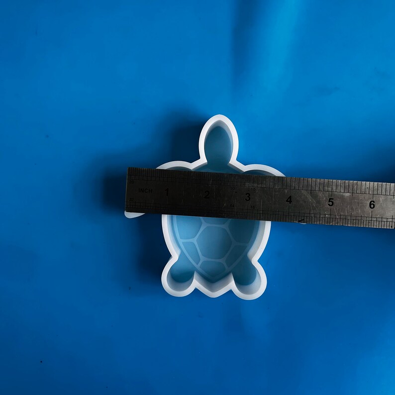 Puede incluir: Un molde blanco con forma de tortuga con una regla que mide su tama&ntilde;o. El molde tiene un dise&ntilde;o detallado de caparaz&oacute;n y mide aproximadamente 7.6 cm de largo. El fondo es de color azul liso. Este molde es adecuado para proyectos de manualidades.