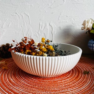 Könnte beinhalten: Weiße Keramikschüssel mit geripptem Design, gefüllt mit getrockneten Blumen in Orange-, Gelb- und Grüntönen. Die Schüssel steht auf einem runden, orangefarbenen Untersetzer.