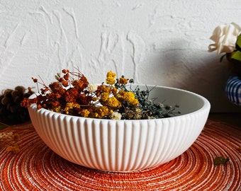 May include: White ceramic bowl with a ribbed design, filled with dried flowers in shades of orange, yellow, and green. The bowl is sitting on a round orange placemat.