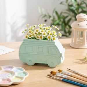 Peut inclure: Un pot de fleurs en forme de camping-car vert clair rempli de marguerites blanches et jaunes. Le pot a des fenêtres rectangulaires et des roues rondes. Des pinceaux et une palette de peinture sont au premier plan.