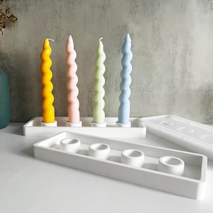 Peut inclure: Un bougeoir en céramique blanche avec quatre puits individuels pour bougies. Le bougeoir est rectangulaire et a une finition lisse et mate. Quatre bougies torsadées colorées sont affichées dans le bougeoir.