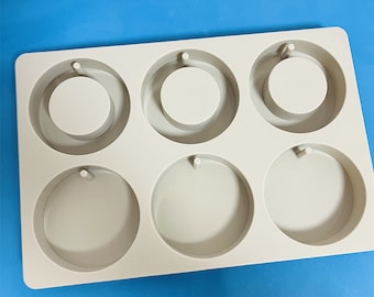Op de afbeelding: Een witte siliconen mal met zes ronde holtes voor het maken van sieraden. Elke holte heeft een klein gaatje bovenaan om het afgewerkte stuk op te hangen.