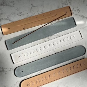 May include: Four incense stick holders made of different materials. The holders are all rectangular and have a design of crescent moons. The holders are in the colours beige, white, grey, and brown.
