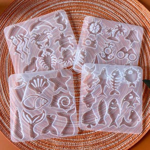May include: Four clear silicone moulds featuring marine-themed designs. The moulds include jellyfish, crabs, seahorses, turtles, seashells, starfish, fish, and nautical elements like anchors and steering wheels. The moulds are on a textured orange surface.