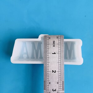 Op de afbeelding: Een doorzichtige siliconen mal met de letters "AM" in een bloklettertype. De mal is ongeveer 2,5 cm breed. De achtergrond is effen lichtblauw.