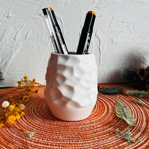 May include: White ceramic pencil holder with a textured surface. Two black markers with orange caps are in the holder. The holder is sitting on a brown and white circular placemat.