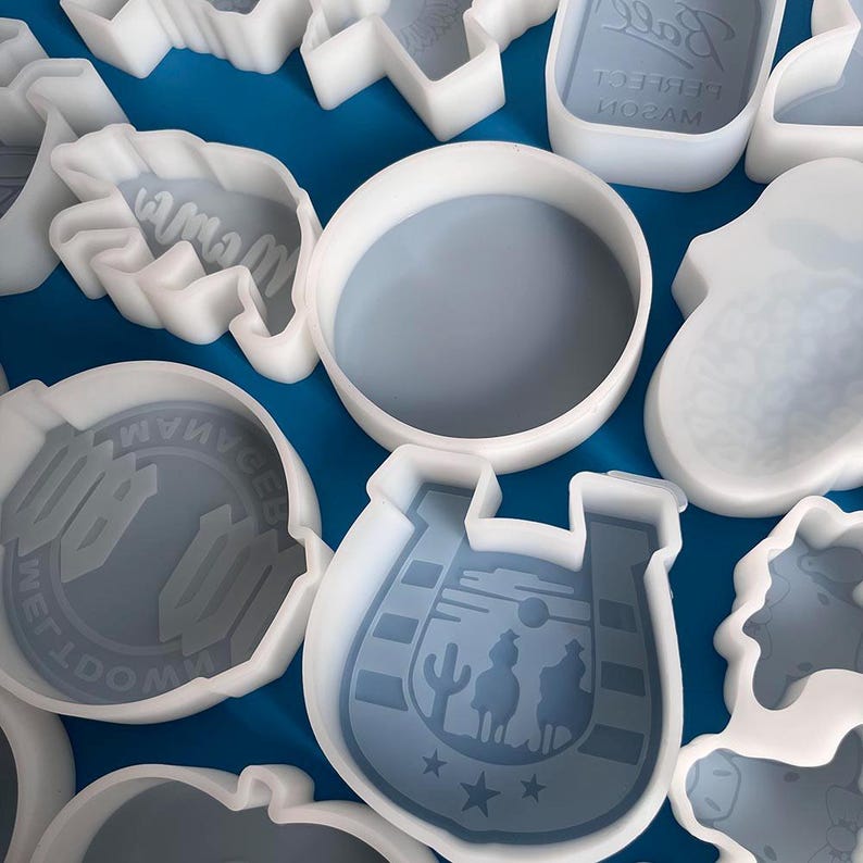May include: A set of white silicone molds for baking. The molds are shaped like a horseshoe, a cowboy hat, a mason jar, a bottle, and a cow. The molds are on a blue surface.