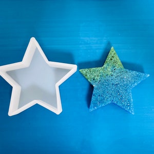Pode incluir: Dois moldes em forma de estrela, um branco e outro azul e verde. O molde branco é de silicone e está vazio. O molde azul e verde é feito de um material texturizado e está cheio de uma substância azul e verde.
