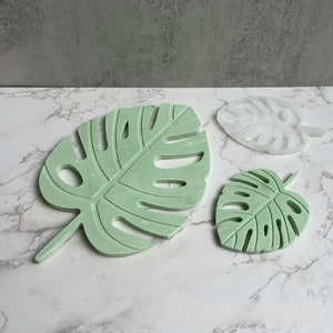 May include: Three green and white monstera leaf-shaped coasters. The coasters are made of a smooth, hard material and have a raised design.