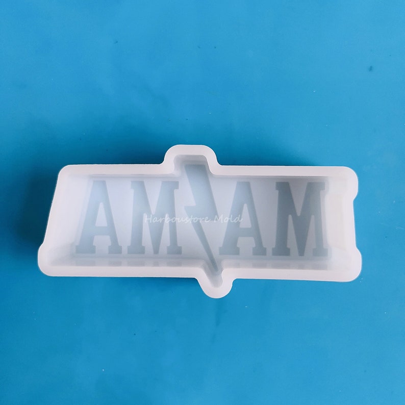 Op de afbeelding: Een witte siliconen mal met de letters "AM AM" in een bloklettertype. Een bliksemvorm staat in het midden. De tekst "Harbourstore Mold" is zichtbaar. De achtergrond is effen blauw.