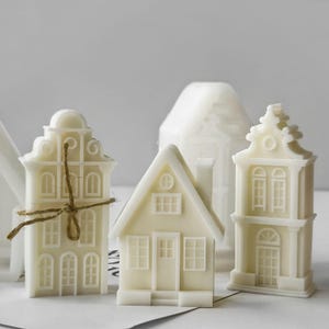 May include: Three off-white wax candles shaped like miniature houses with detailed architectural features including windows, doors, and gables. One candle is tied with a thin brown twine string, while the others stand independently on a flat surface.